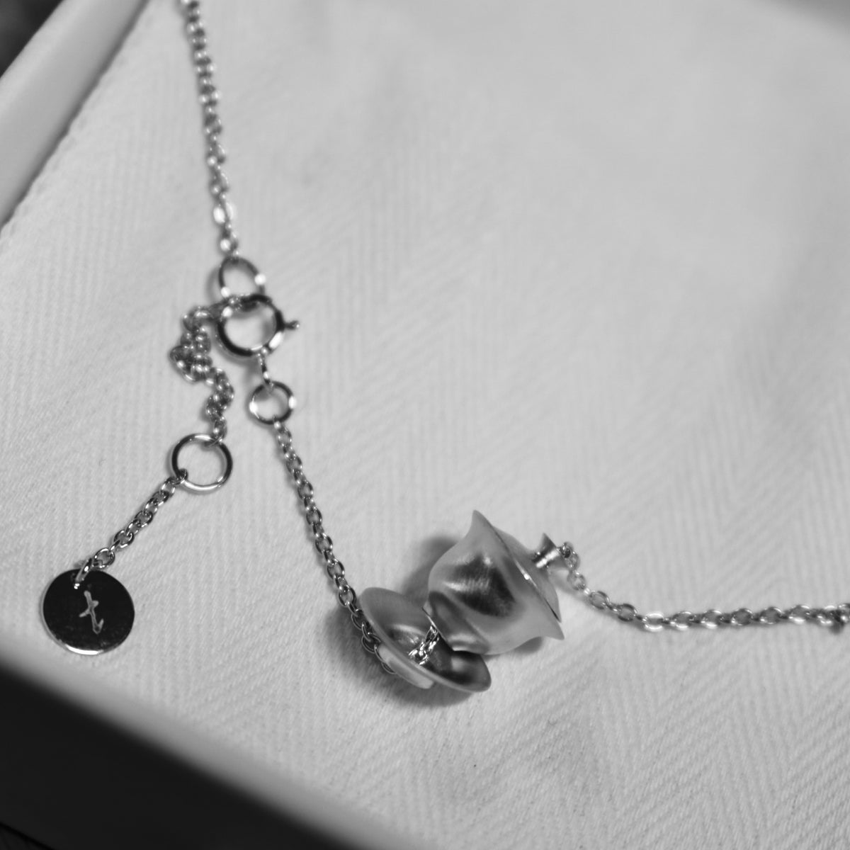 A black-and-white close-up of a silver Gai Wan-shaped necklace pendant on fabric.