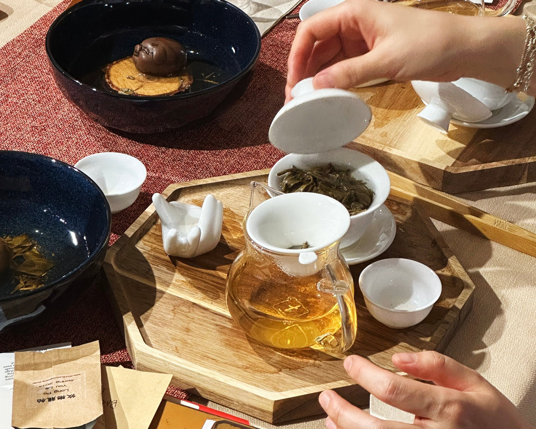 Hands pouring brewed tea into a gaiwan surrounded by tasting bowls and tea leaves.