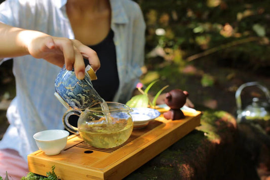 Hand pouring tea from a blue-and-white teapot into a glass pitcher outdoors near a small clay tea pet