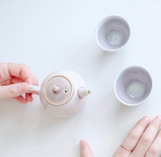 Overhead view of hands opening a small white teapot lid beside two empty teacups.