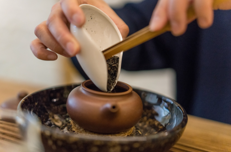 Hands pouring dark tea leaves from a white gaiwan into a small clay teapot