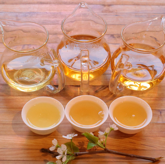 Three glass pitchers and three cups of steeped tea in varying gold shades, with a flowering branch.