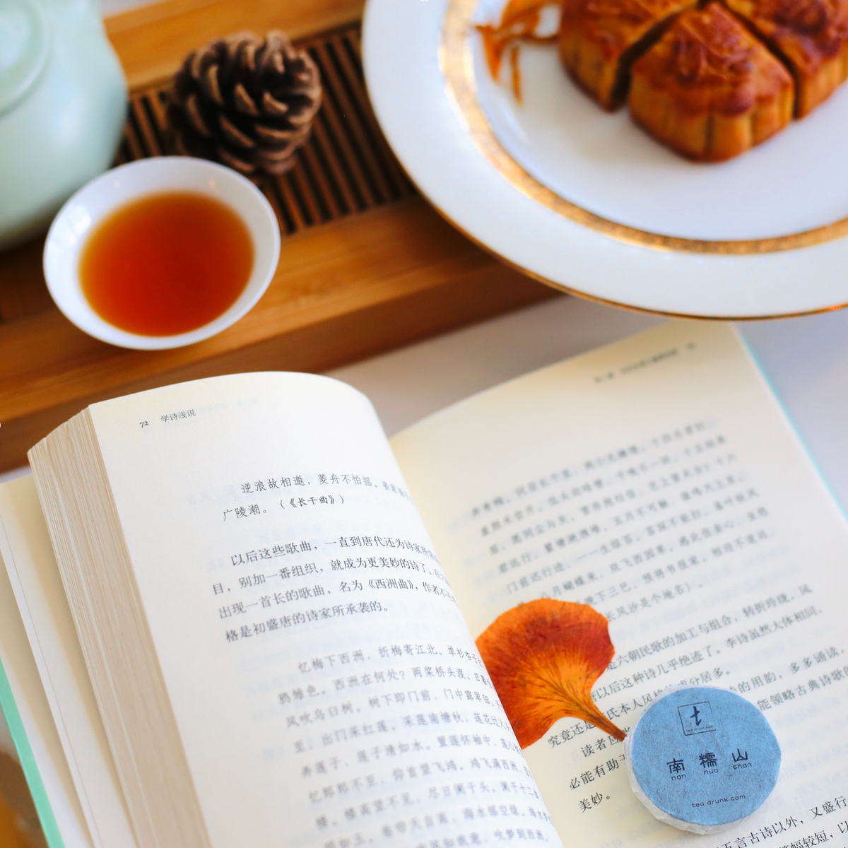 An open book with a ginkgo leaf bookmark beside a cup of tea, mooncakes, and a compressed tea cake.