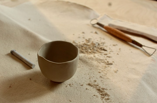 A handmade ceramic teacup being shaped on a pottery table with carving tools.
