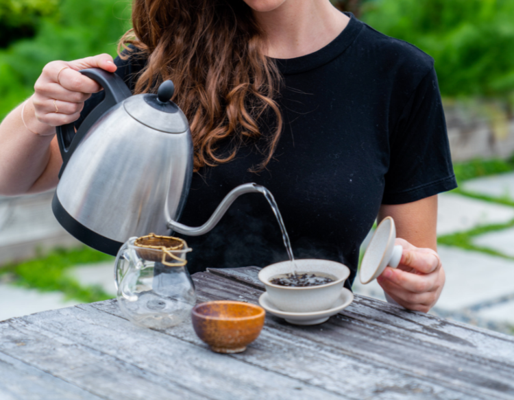 Woman pouring hot water from a gooseneck kettle into a gaiwan of tea leaves