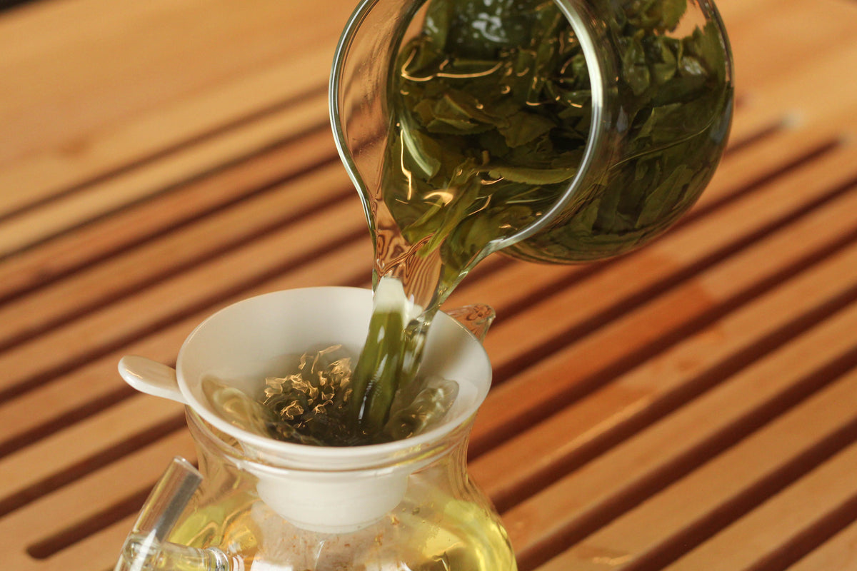 A handheld tea strainer filtering brewed tea into a fairness pitcher.