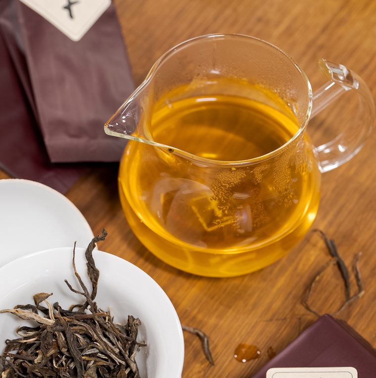 Amber tea in a clear glass fairness pitcher beside loose tea leaves.